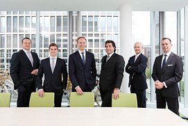 Schönes Teamfoto einer Gruppe von Geschäftsleuten in Anzügen vor einer Glasfassade für ein Unternehmen aus Bonn.